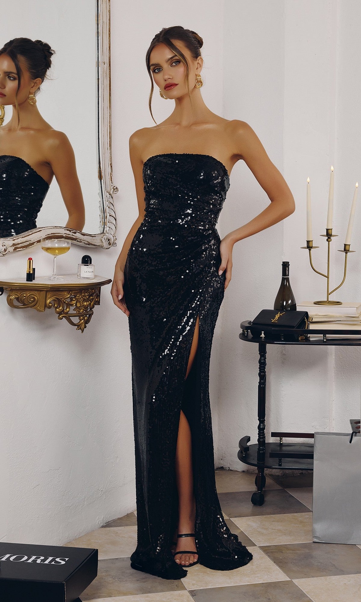 A woman in a black Long Prom Dress: Amoris T724 stands indoors by a mirror and a table with candles, wine, and a glass.